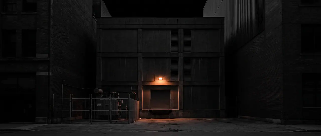 Windowless concrete facade of a data centre at night, wedged between taller neighbouring buildings; the only light source is a copper-coloured lamp above the loading dock, next to a fenced transformer yard.