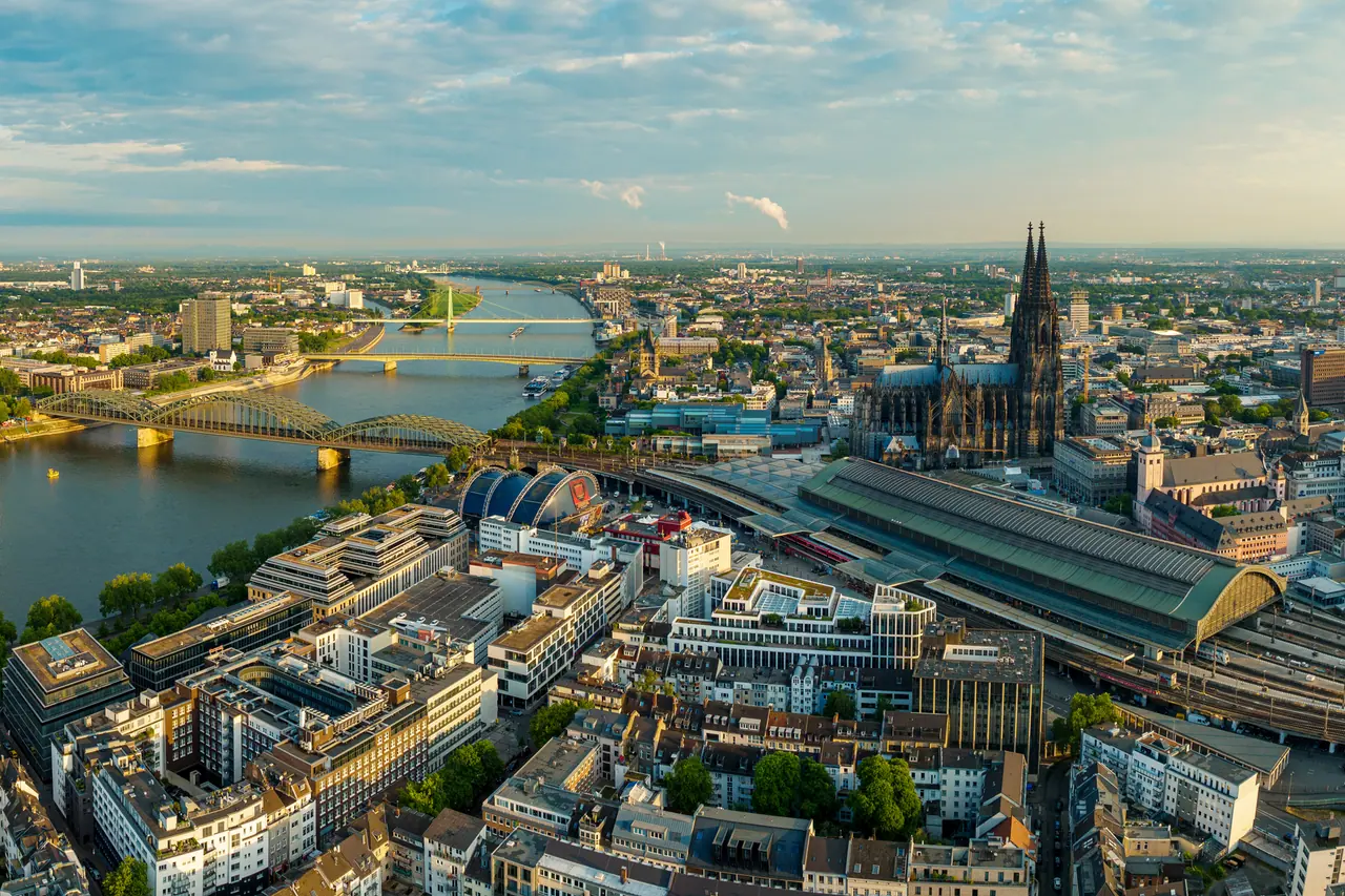 Luftaufnahme von Köln bei tief stehender Sonne: links der Rhein mit der Hohenzollernbrücke und zwei weiteren Brücken, rechts der Dom, davor die Halle des Hauptbahnhofs und die Dächer der Innenstadt.