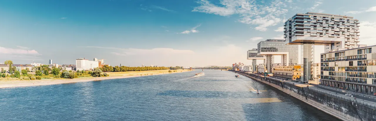 Der Rhein in Köln an einem klaren Sommertag: rechts am Ufer die drei Kranhäuser des Rheinauhafens, links die Wiesen und Bäume der gegenüberliegenden Rheinseite, auf dem Fluss zwei Schiffe.
