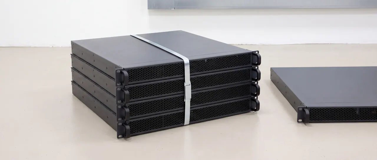 Four identical anthracite-coloured rack chassis lie flush on top of each other on the light screed of an empty technical area. A tensioned bare steel strapping band runs horizontally around the block, visibly pressing into the edges of the chassis and closed at the front by a flat-pressed seal; it is the brightest line in the picture. To the right of it, a fifth, exactly identical chassis lies individually and unbound on the floor.