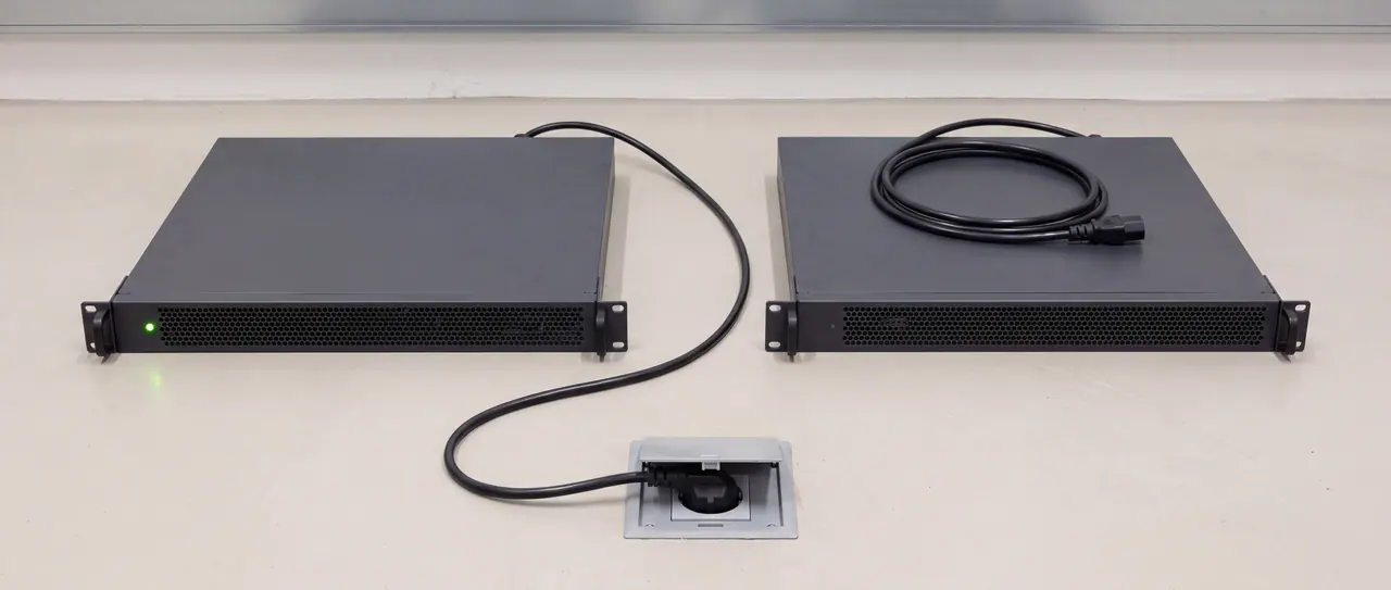 Two identical anthracite-coloured rack chassis stand side by side on the light screed of an empty technical area, both with the same perforated honeycomb front and the same rack ears. On the left one, the black IEC cable runs in a curve across the floor into a flush-mounted connection socket and is plugged in there; a single small green dot lights up on its front. On the right one, the same cable lies as a flat coiled ring on the cover, the plug free, the front unlit.