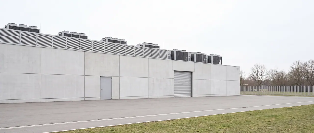 Long side of a windowless technical building made of light precast concrete parts under an overcast sky: a steel door and a roller shutter in the facade, on the roof a row of identical dry coolers behind a louvre blind, in front of it an empty asphalt driveway.