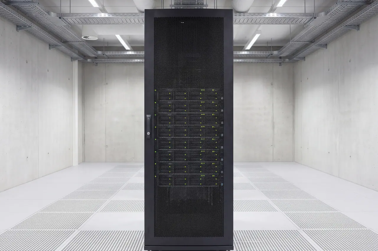 Einzelner Serverschrank von vorn, die Lochblechtür geschlossen und mit einem bündigen Schwenkgriff samt Schließzylinder gesichert. Durch die gleichmäßige Lochung sind die baugleichen Gerätefronten als regelmäßiger Stapel zu sehen, jede mit ihrem eigenen grünen Statuslicht.