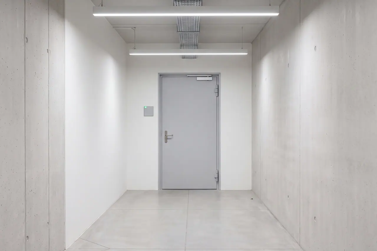 Zutrittstür am Ende eines Betongangs: ein graues Stahlblatt in einer Stahlzarge mit sichtbaren Bändern, Drückergarnitur und obenliegendem Türschließer. Daneben auf Handhöhe ein schmuckloses Lesegerät mit einem einzelnen grünen Punkt; der Gang ist leer und durchgehend beleuchtet.