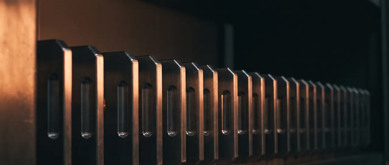 Long row of uniform brushed steel front panels, seen flat from the left; each panel has a milled recessed grip that catches the light as a sharp edge.
