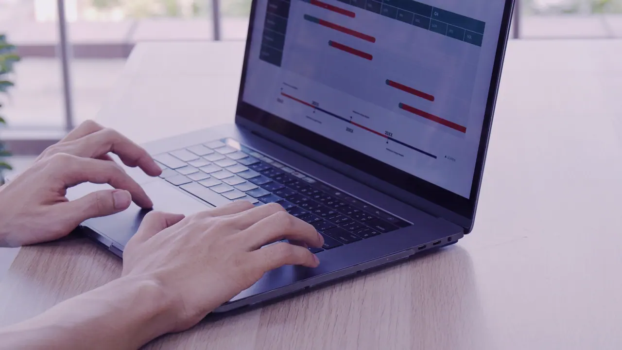 Two hands on the keyboard of an open laptop on a light wooden table by the window; on the screen a timeline with red bars.