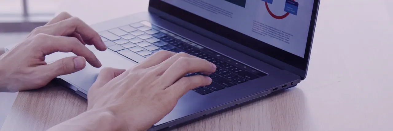 Two hands typing on the keyboard of a laptop on a light table; on the screen behind it a chart and text blocks.