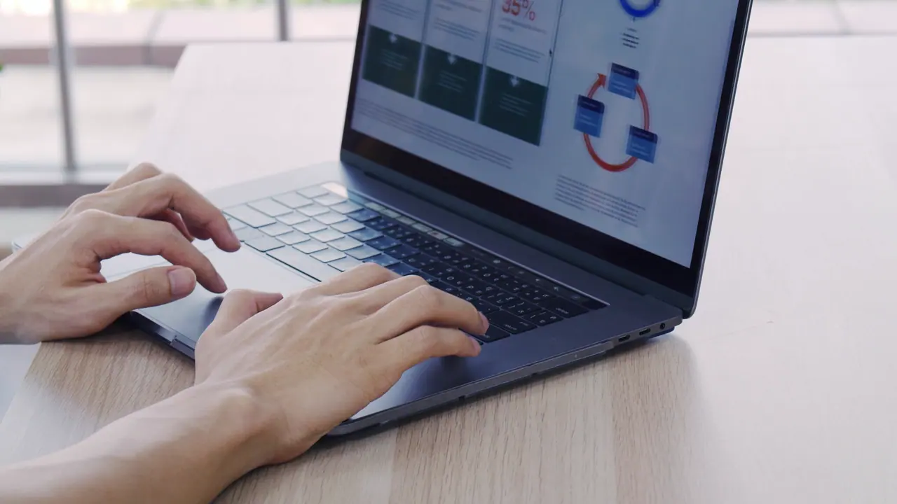 Hands on the keyboard of an open laptop on a light wooden table; the screen shows an overview with bars and charts.